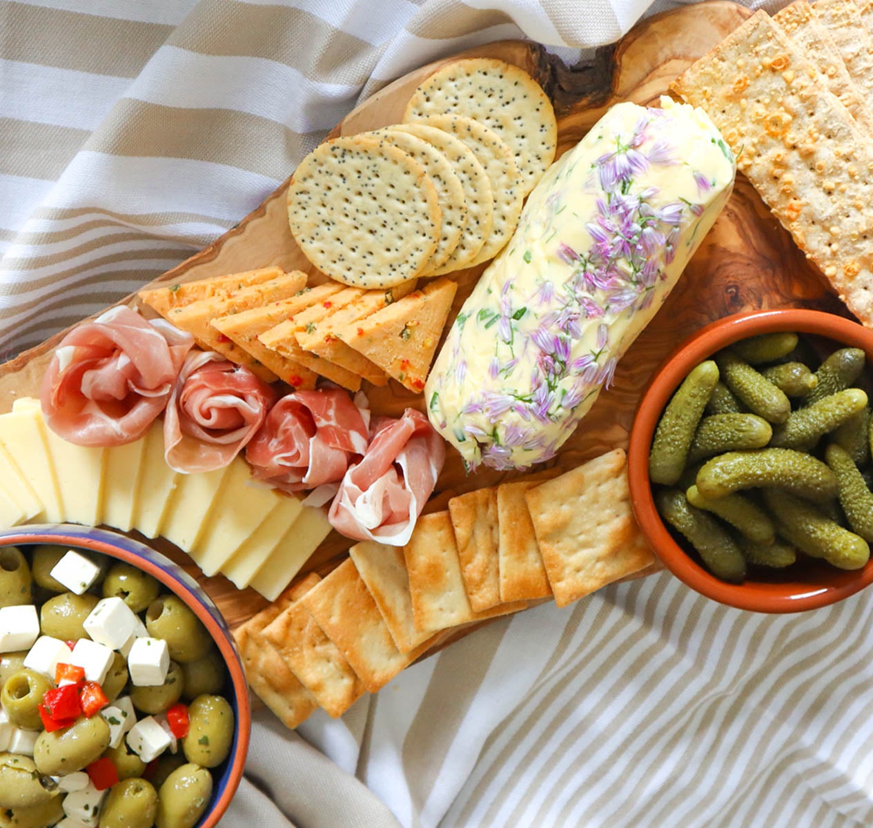 Hausgemachte Schnittlauchblütenbutter A wooden platter featuring homemade chive flower butter, assorted cheeses, crackers, olives, and pickles.