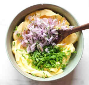 A bowl of butter mixed with chive flowers and chopped chives, with a wooden spoon resting inside.