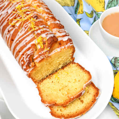 Sliced lemon drizzle cake with icing and lemon zest on top, served with a cup of tea.
