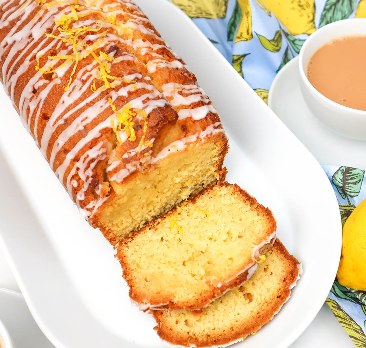 Sliced lemon drizzle cake with icing and lemon zest on top, served with a cup of tea.