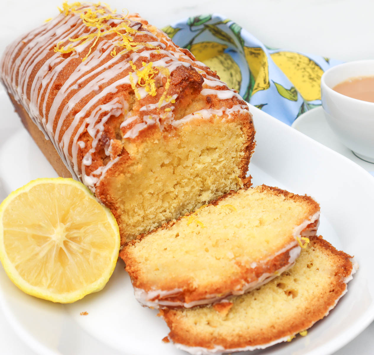 Lemon Drizzle Cake A sliced lemon drizzle cake with a lemon slice and a cup of tea in the background.