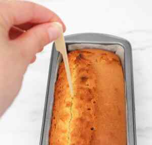 A hand inserting a toothpick into a freshly baked lemon drizzle cake in a loaf pan.