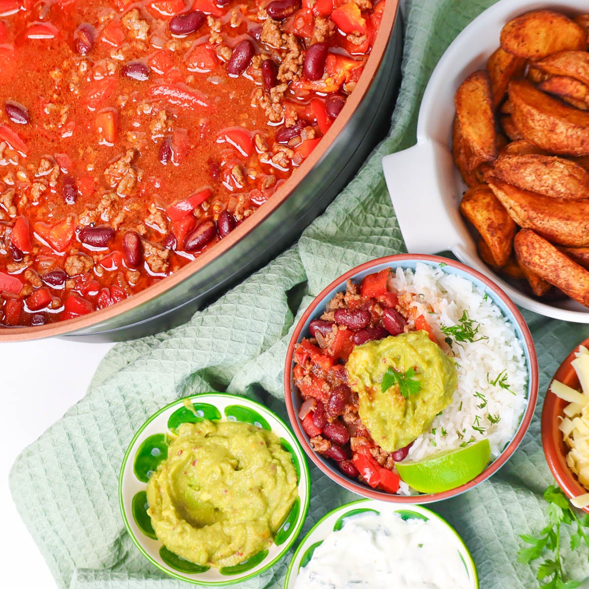 Slow Cooker Chili Con Carne A bowl of chili con carne with rice, topped with guacamole and lime, alongside sides of guacamole and sour cream.