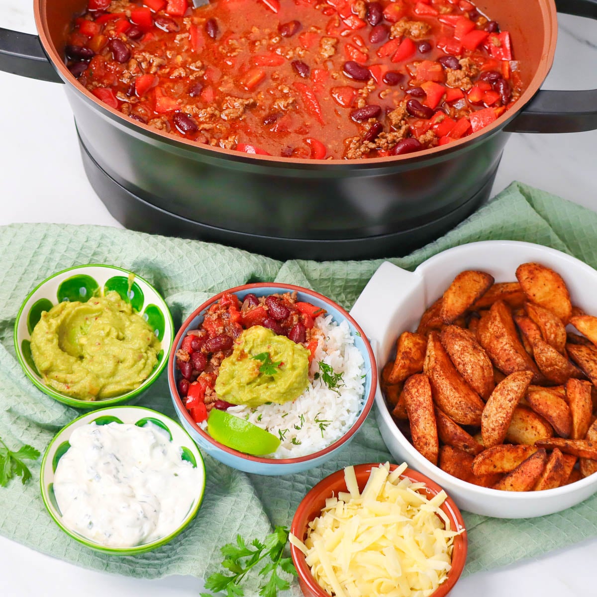Slow Cooker Chili Con Carne A bowl of chili con carne with toppings, served with sides of guacamole, sour cream, and seasoned potato wedges.