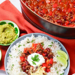 A bowl of chili con carne served over rice with lime wedges and guacamole on the side.