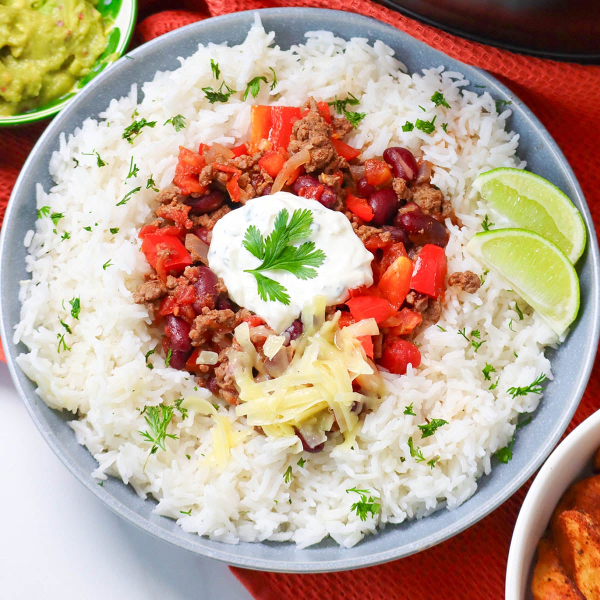 Slow Cooker Chili Con Carne A bowl of slow cooker chili con carne served over rice, garnished with sour cream, cheese, and lime wedges.