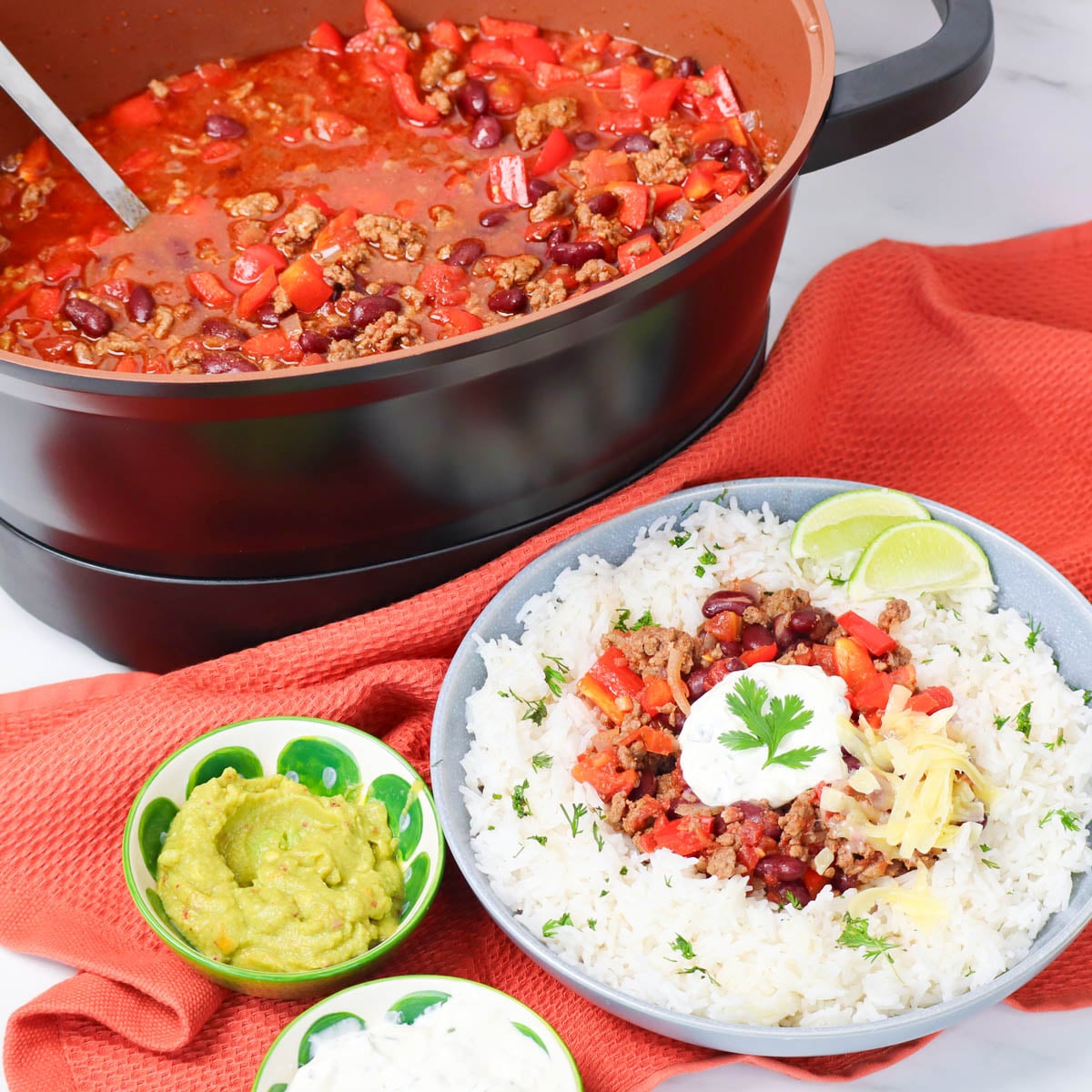 Slow Cooker Chili Con Carne A bowl of chili con carne served over rice, garnished with cilantro, cheese, and lime, alongside guacamole and sour cream.