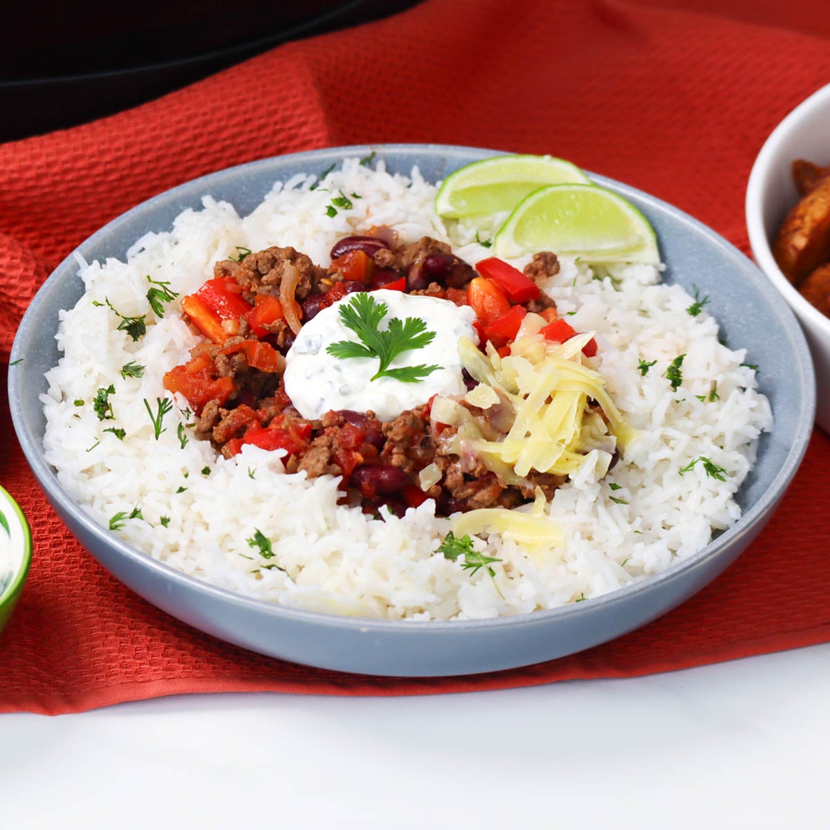 Slow Cooker Chili Con Carne A bowl of slow cooker chili con carne served over rice, garnished with cilantro, sour cream, and lime slices.