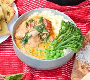 A bowl of creamy chicken with spinach, served with mashed potatoes and green vegetables on a red striped cloth.