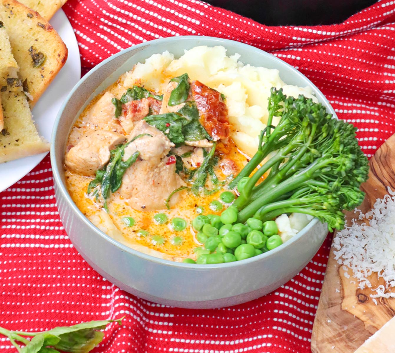 Slow Cooker Marry Me Chicken A bowl of creamy chicken with spinach, served with mashed potatoes and green vegetables on a red striped cloth.