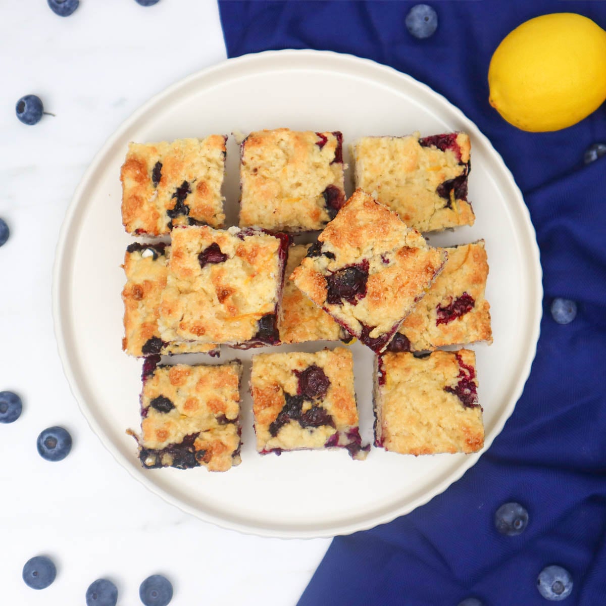 American Style Blueberry Biscuits A plate of golden-brown blueberry biscuits cut into squares, surrounded by fresh blueberries and a lemon.