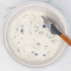 A bowl of biscuit dough mixed with blueberries and a spatula resting inside.