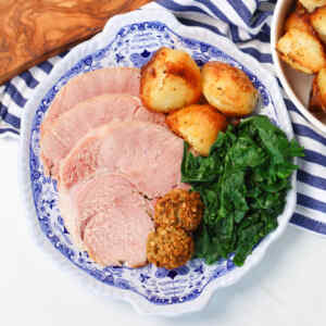 Sliced slow cooker ham served with roasted potatoes, greens, and stuffing balls on a decorative plate.