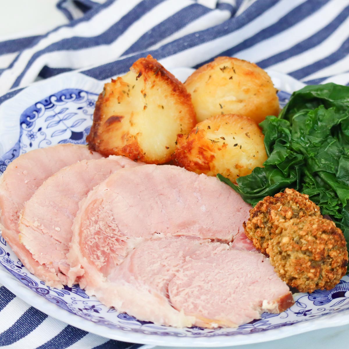 Slow Cooker Ham Sliced slow cooker ham served with crispy potatoes, greens, and stuffing balls on a decorative plate.